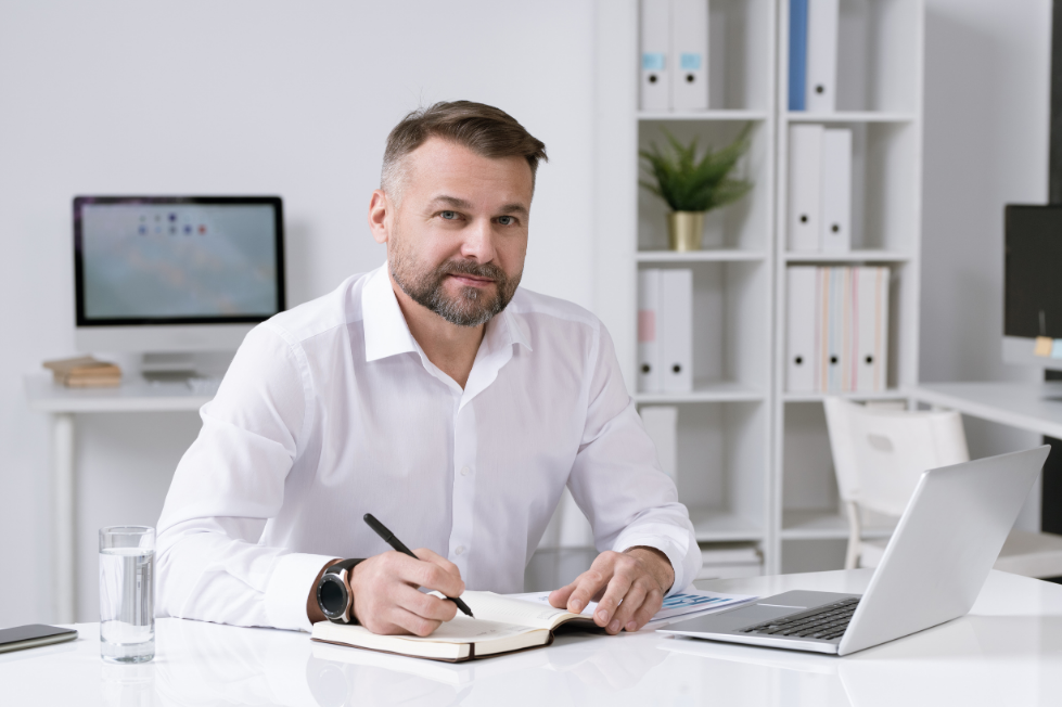 A professional man at a modern office desk reflecting and writing in a notebook next to a laptop while receiving non-clinical supervision